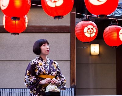 Woman in a floral kimono gazes at red Japanese lanterns outside a traditional building, creating a warm, contemplative festival mood.