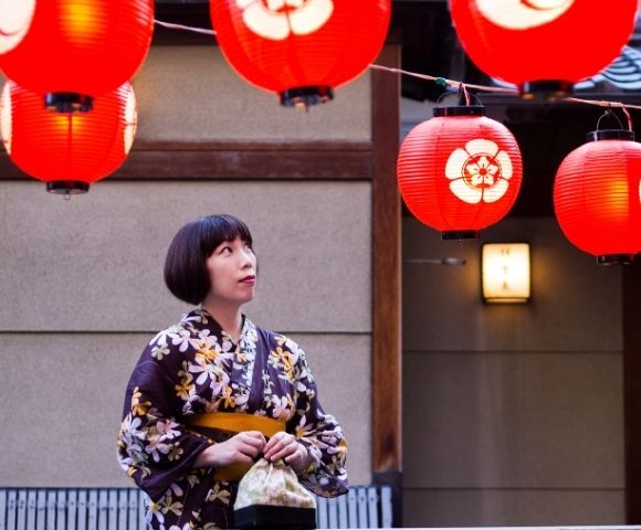 Woman in a floral kimono gazes at red Japanese lanterns outside a traditional building, creating a warm, contemplative festival mood.