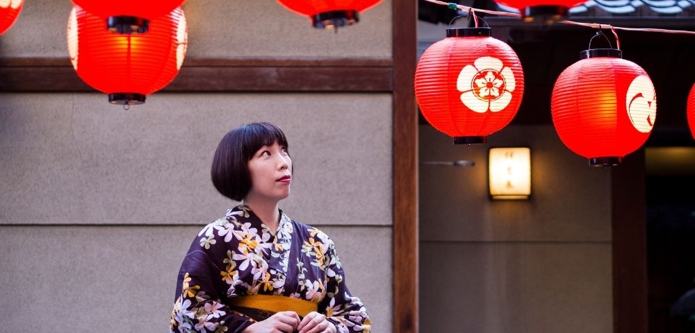 Woman in a floral kimono gazes at red Japanese lanterns outside a traditional building, creating a warm, contemplative festival mood.