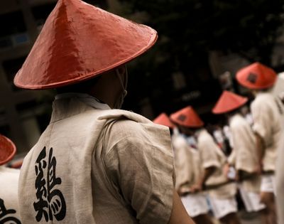A solemn procession of people in cream robes and red conical hats moves through a dim street, with a large black Chinese character on the nearest robe.
