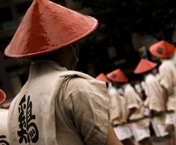 A solemn procession of people in cream robes and red conical hats moves through a dim street, with a large black Chinese character on the nearest robe.