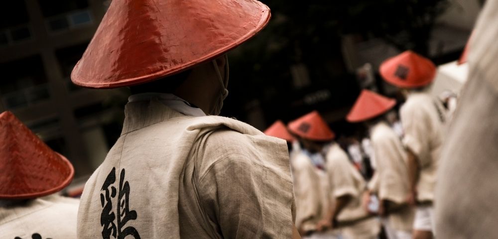 A solemn procession of people in cream robes and red conical hats moves through a dim street, with a large black Chinese character on the nearest robe.