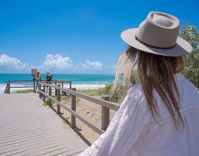 Woman in a wide-brimmed hat and white shirt walks along a beach boardwalk toward turquoise ocean under a bright, cloud-dotted sky.