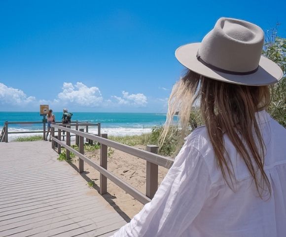 Woman in a wide-brimmed hat and white shirt walks along a beach boardwalk toward turquoise ocean under a bright, cloud-dotted sky.
