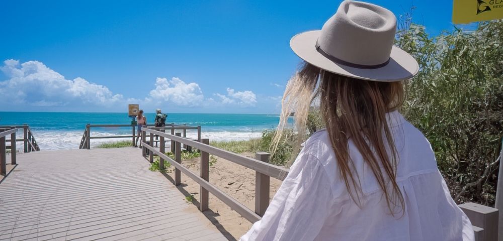 Woman in a wide-brimmed hat and white shirt walks along a beach boardwalk toward turquoise ocean under a bright, cloud-dotted sky.