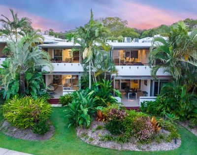 Two-story tropical resort surrounded by lush palms and landscaped gardens, glowing under a pink-and-blue sunset sky; serene, inviting atmosphere.