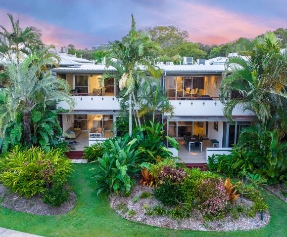 Two-story tropical resort surrounded by lush palms and landscaped gardens, glowing under a pink-and-blue sunset sky; serene, inviting atmosphere.
