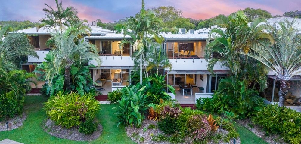 Two-story tropical resort surrounded by lush palms and landscaped gardens, glowing under a pink-and-blue sunset sky; serene, inviting atmosphere.