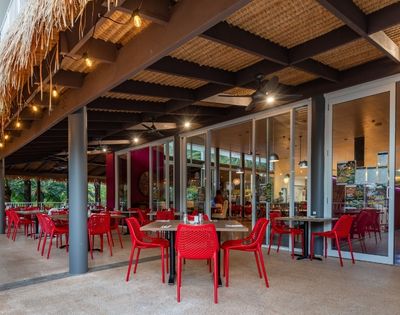 Open-air tropical restaurant patio with red tables and chairs, palm trees, thatched roof, warm lights, and inviting indoor dining area.