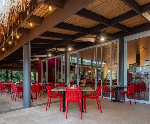 Open-air tropical restaurant patio with red tables and chairs, palm trees, thatched roof, warm lights, and inviting indoor dining area.