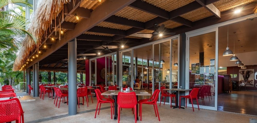 Open-air tropical restaurant patio with red tables and chairs, palm trees, thatched roof, warm lights, and inviting indoor dining area.