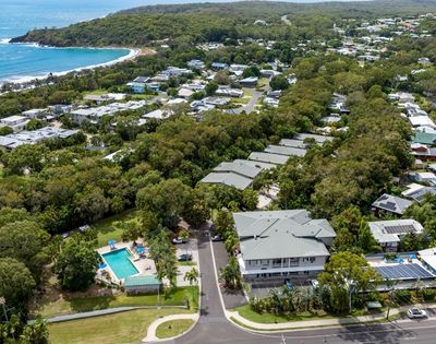 Aerial view of a leafy coastal suburb with homes, roads, and a turquoise swimming pool near a curved beach and blue ocean, creating a peaceful scene.
