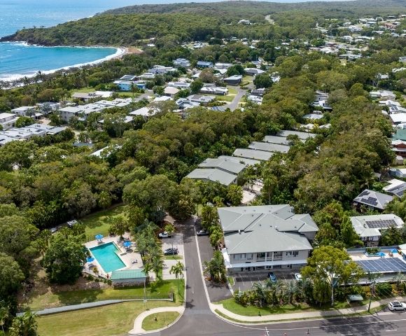 Aerial view of a leafy coastal suburb with homes, roads, and a turquoise swimming pool near a curved beach and blue ocean, creating a peaceful scene.