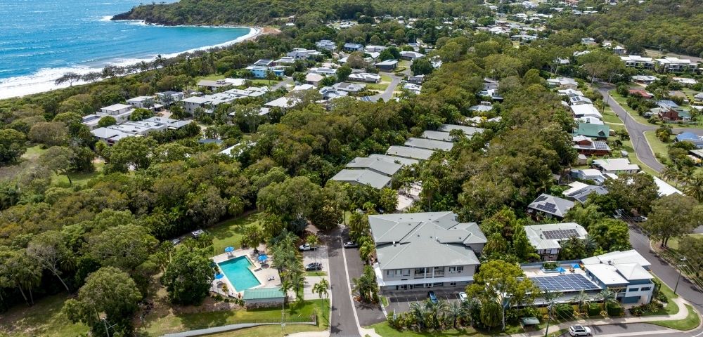 Aerial view of a leafy coastal suburb with homes, roads, and a turquoise swimming pool near a curved beach and blue ocean, creating a peaceful scene.