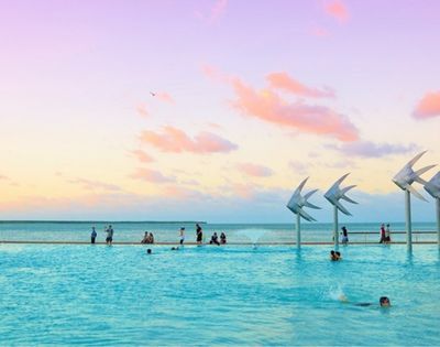 Turquoise infinity pool meets the ocean beneath a pastel sunset, with swimmers, waders, and sail-like sculptures creating a serene scene.