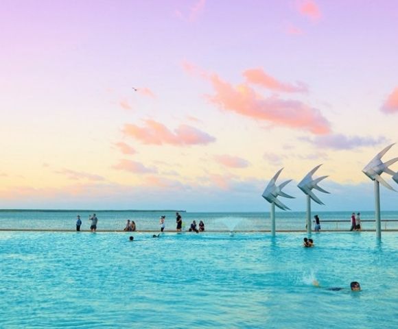 Turquoise infinity pool meets the ocean beneath a pastel sunset, with swimmers, waders, and sail-like sculptures creating a serene scene.