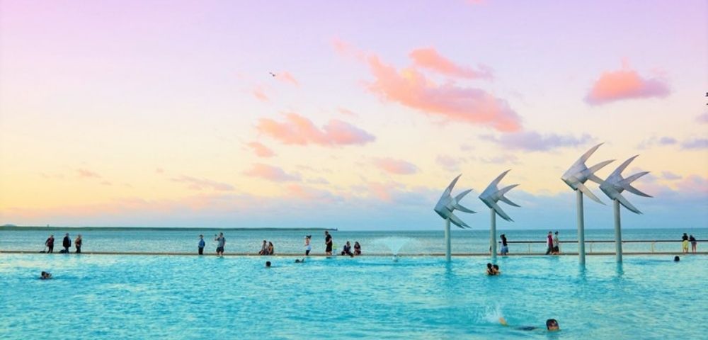 Turquoise infinity pool meets the ocean beneath a pastel sunset, with swimmers, waders, and sail-like sculptures creating a serene scene.