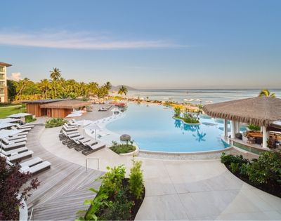 A luxurious resort pool with pristine blue water, loungers, and a thatched-roof bar faces a serene ocean under a clear blue sky, with palm trees lining the sandy beach.