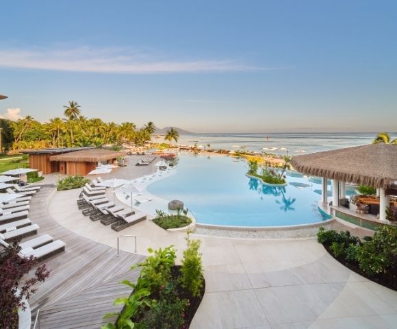 A luxurious resort pool with pristine blue water, loungers, and a thatched-roof bar faces a serene ocean under a clear blue sky, with palm trees lining the sandy beach.
