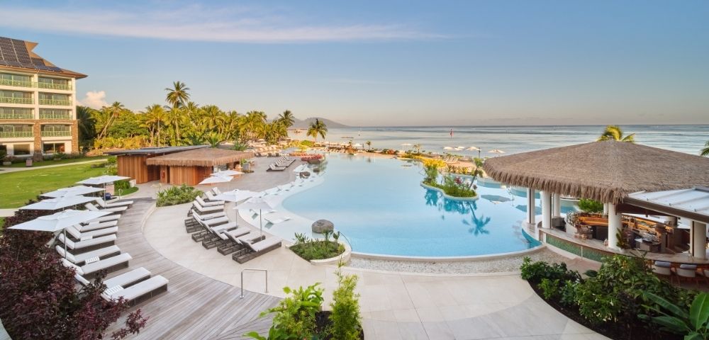 A luxurious resort pool with pristine blue water, loungers, and a thatched-roof bar faces a serene ocean under a clear blue sky, with palm trees lining the sandy beach.