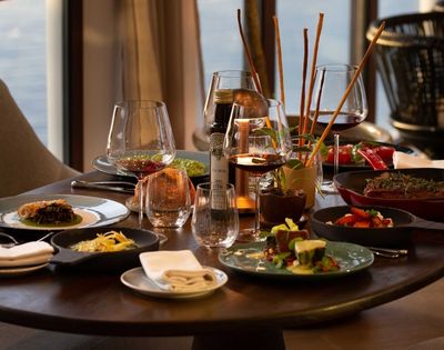 Elegant restaurant table set with wine glasses, cocktails, shared gourmet dishes, candles, and warm sunset light creating an inviting mood.