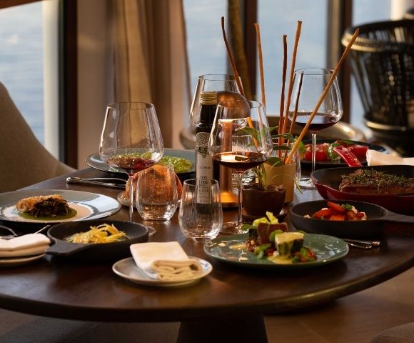 Elegant restaurant table set with wine glasses, cocktails, shared gourmet dishes, candles, and warm sunset light creating an inviting mood.