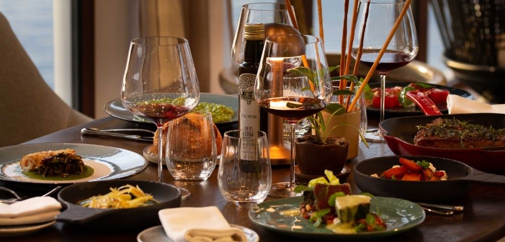 Elegant restaurant table set with wine glasses, cocktails, shared gourmet dishes, candles, and warm sunset light creating an inviting mood.