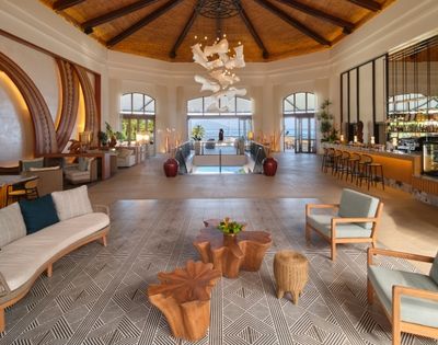 Elegant luxury hotel lobby with a vaulted wood ceiling, sculptural chandelier, lounge seating, patterned carpet, bar, and sunlit pool view.