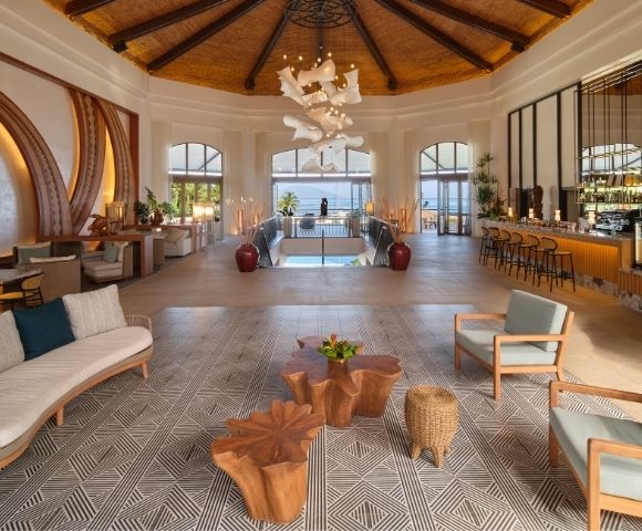 Elegant luxury hotel lobby with a vaulted wood ceiling, sculptural chandelier, lounge seating, patterned carpet, bar, and sunlit pool view.