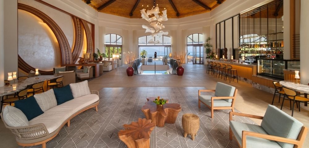 Elegant luxury hotel lobby with a vaulted wood ceiling, sculptural chandelier, lounge seating, patterned carpet, bar, and sunlit pool view.