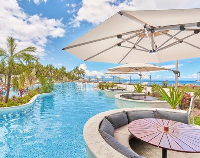 Resort pool curves past palm trees and ocean views, with shaded umbrellas, lounge seating, and a round table creating a serene tropical retreat.