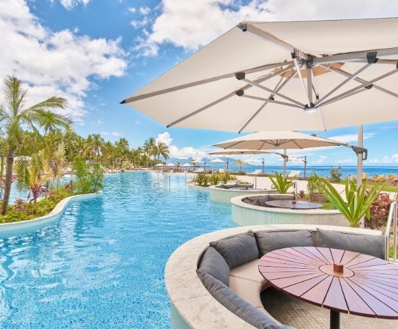 Resort pool curves past palm trees and ocean views, with shaded umbrellas, lounge seating, and a round table creating a serene tropical retreat.