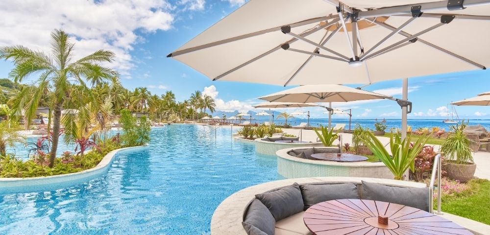 Resort pool curves past palm trees and ocean views, with shaded umbrellas, lounge seating, and a round table creating a serene tropical retreat.