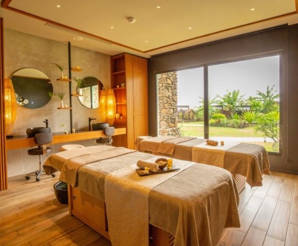 Serene luxury spa treatment room with two massage beds, warm lighting, stone accents, round mirrors, and large windows overlooking a lush garden.