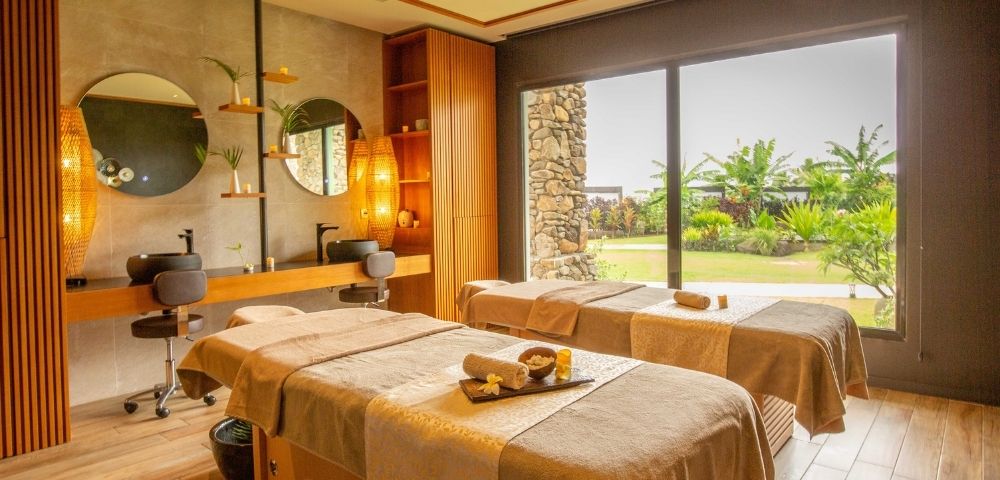 Serene luxury spa treatment room with two massage beds, warm lighting, stone accents, round mirrors, and large windows overlooking a lush garden.
