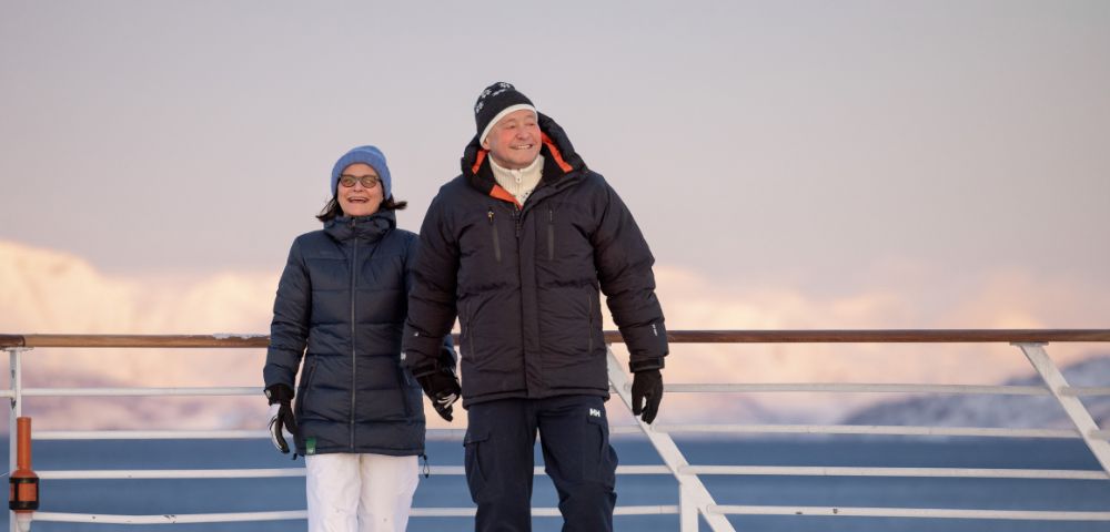 Two people bundled in winter jackets and hats stand on the deck of a ship, looking out at a calm, blue sea under a pastel sky, with snowy mountains in the distance.