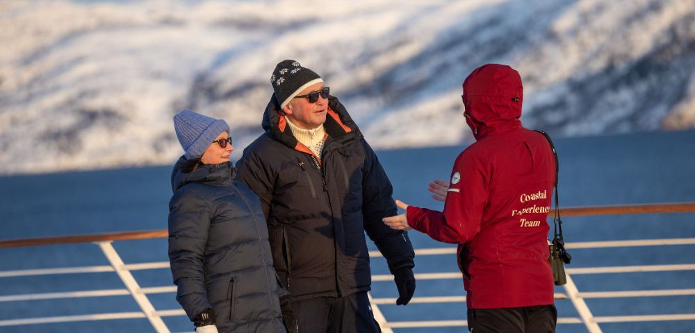 Three people in winter clothing stand on a deck railing with a snow-covered mountain range and blue water in the background. A person in a red jacket with 