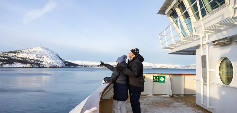 Two people in winter clothing stand on the deck of a ship, looking out at a snow-covered mountain range and a calm body of water under a clear sky.
