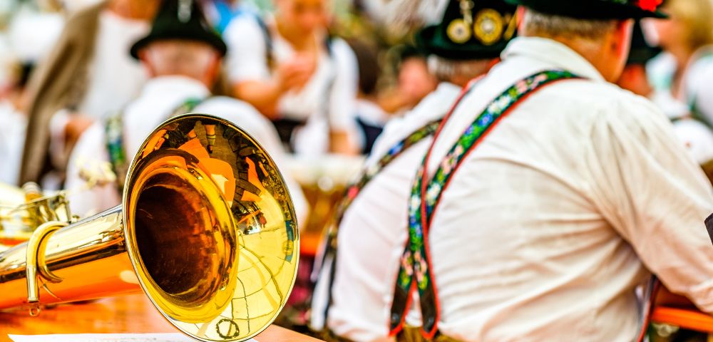 A close-up of a shiny brass tuba bell sits on a wooden table, reflecting a festive, blurred background of people in traditional Bavarian attire.