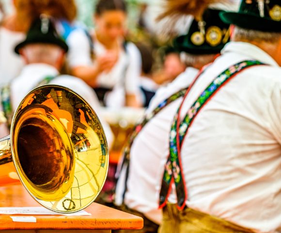 A close-up of a shiny brass tuba bell sits on a wooden table, reflecting a festive, blurred background of people in traditional Bavarian attire.