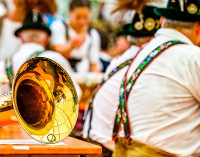 A close-up of a shiny brass tuba bell sits on a wooden table, reflecting a festive, blurred background of people in traditional Bavarian attire.