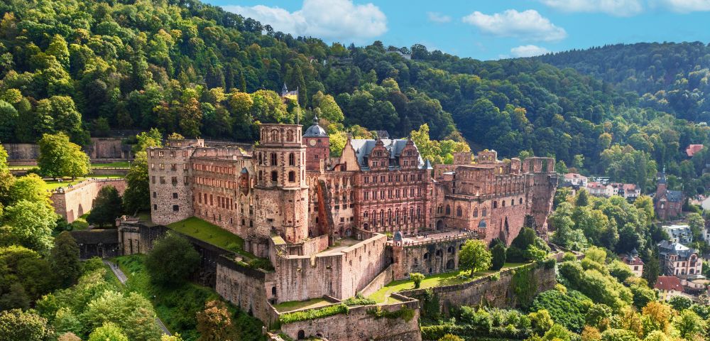 Heidelberg Castle, a historic fortress, perched on a hillside overlooking a town, surrounded by lush green trees under a bright blue sky with scattered clouds.