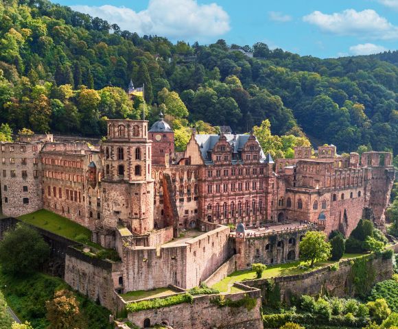 Heidelberg Castle, a historic fortress, perched on a hillside overlooking a town, surrounded by lush green trees under a bright blue sky with scattered clouds.
