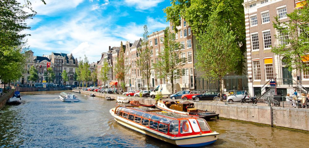 A canal in Amsterdam with a tour boat cruising past charming, narrow buildings under a bright blue, cloudy sky.
