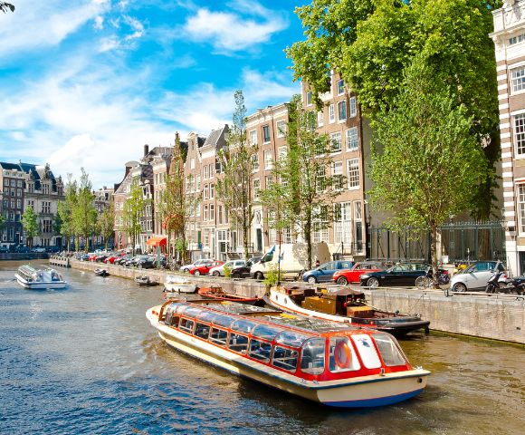 A canal in Amsterdam with a tour boat cruising past charming, narrow buildings under a bright blue, cloudy sky.
