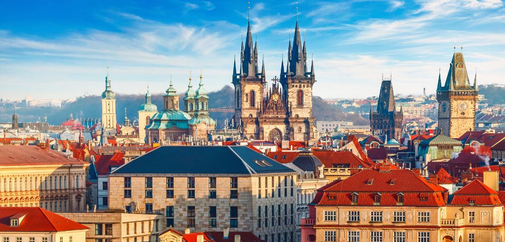 Panoramic Prague skyline featuring the Gothic Church of Our Lady before Týn, red rooftops, Old Town buildings, and a bright blue sky.