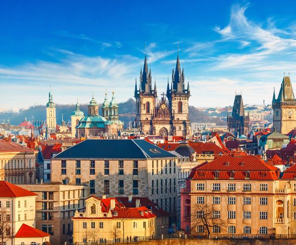 Panoramic Prague skyline featuring the Gothic Church of Our Lady before Týn, red rooftops, Old Town buildings, and a bright blue sky.