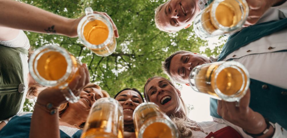 Friends smile and raise glasses of beer toward the camera in a low-angle outdoor toast, creating a joyful, celebratory mood beneath leafy trees.