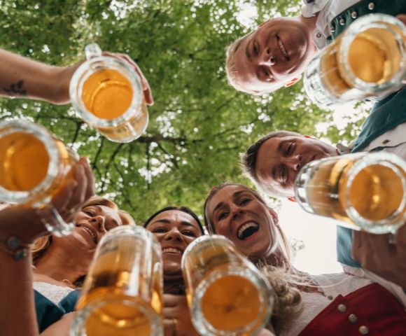Friends smile and raise glasses of beer toward the camera in a low-angle outdoor toast, creating a joyful, celebratory mood beneath leafy trees.