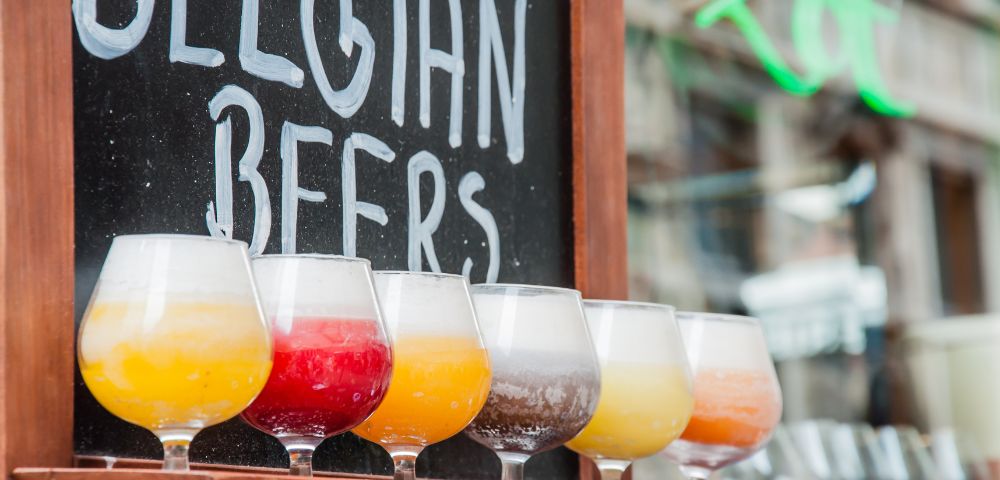 Colorful Belgian beers in tasting glasses line a bar beneath a chalkboard reading “Belgian Beers,” creating a lively, inviting pub atmosphere.